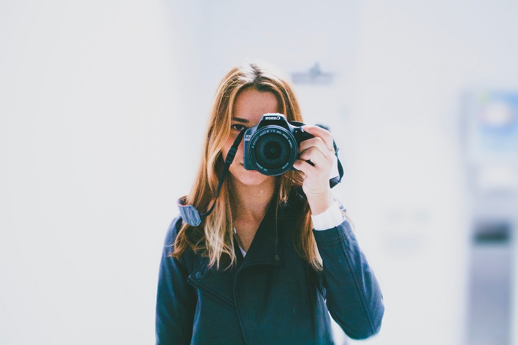 Foto de menina branca e loira com uma câmera na mão e fundo branco.