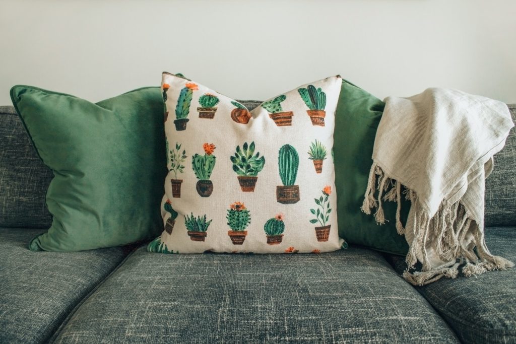 Foto de um sofá com três almofadas verdes e brancas com imagens de cactos na do meio para deixar a decoração mais agradável.