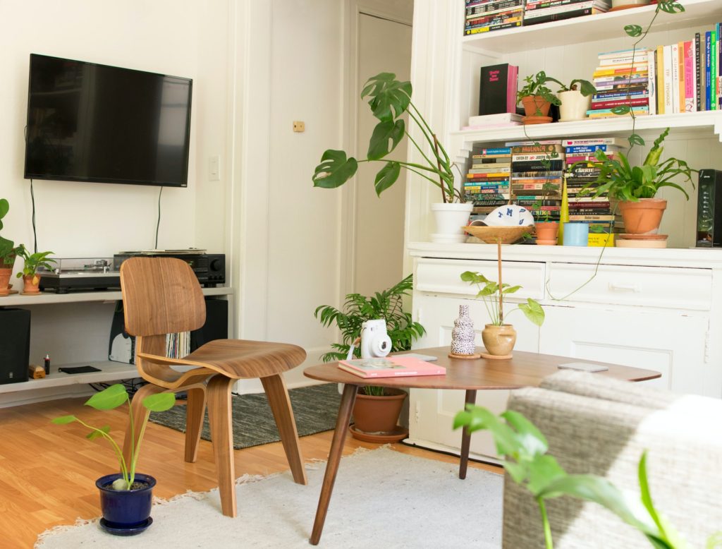 Foto mostrando a decoração de um sala, parece apenas a pontinha do sofá cinza, uma mesinha de centro de madeira com uma cadeira de madeira ao lado e de fundo há uma TV e uma estante embutida com livros.