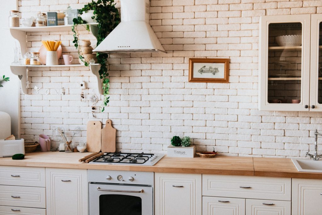 Imagem mostra um papel de parede de tijolinhos na decoração de uma cozinha e ao redor há itens típicos do ambiente como fogão, armário e prateleiras.