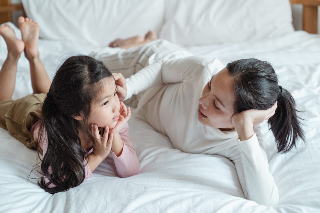 Uma mulher asiática conversa sobre diversidade com uma criança enquanto estão deitadas em uma cama.