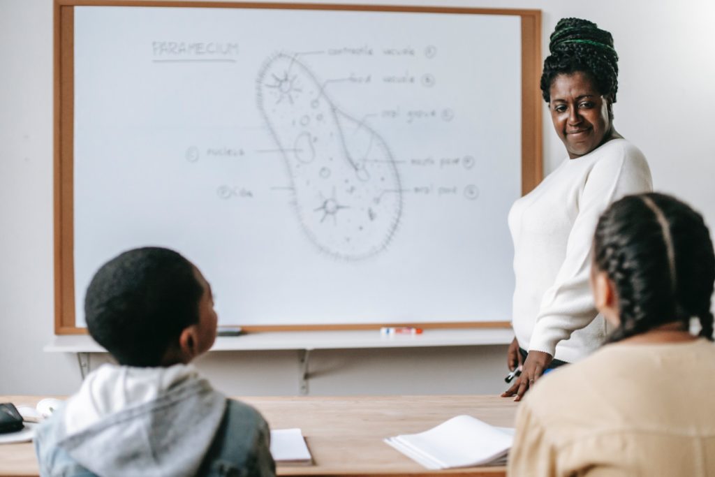 Imagem representa a metodologia pedagógica tradicional com uma professora negra ao lado de um quadro branco em frente a dois alunos também negros, um menino e uma menina.