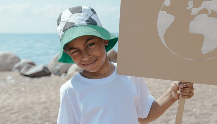 Criança negra com chapéu de praia e camiseta branca segura um cartaz de papelão com imagem do planeta na praia.
