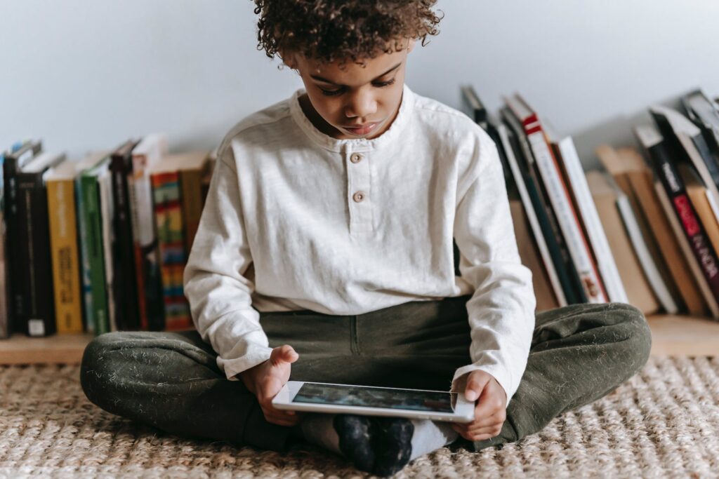 Criança negra segurando um tablet e olhando pra baixo enquanto está sentado na frente de alguns livros