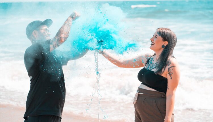 Casal de um homem e uma mulher, brancos e com tatuagens no corpo, seguram objeto que faz fumaça azul em uma praia, em um chá revelação.